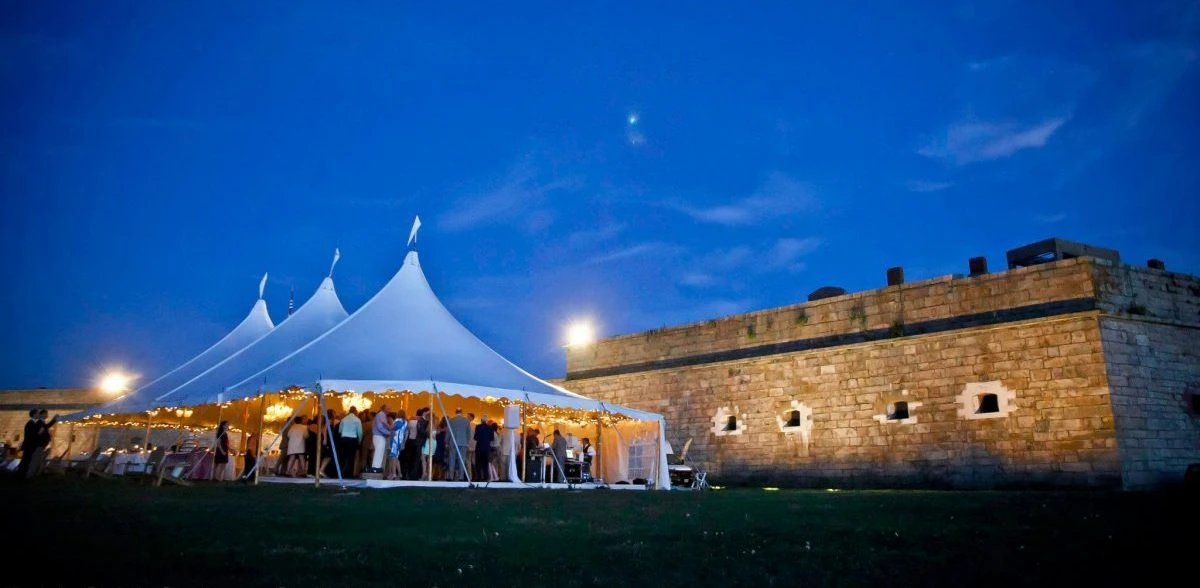 An illuminated white wedding tent beside the stone walls of Fort Adams at dusk
